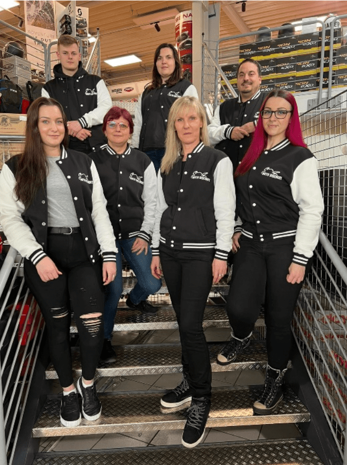 Das Team von Moto Discount GmbH posiert in einheitlicher Motorradbekleidung auf einer Treppe in einem Verkaufsraum.
